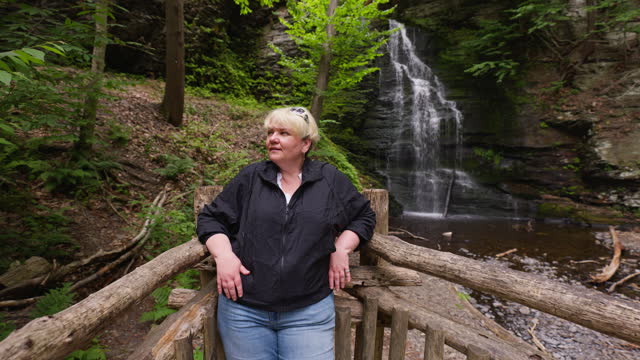 Smiling blonde woman, tourist posing near waterfalls in the forest of Bushkill Falls, PA. Footage with pull camera motion