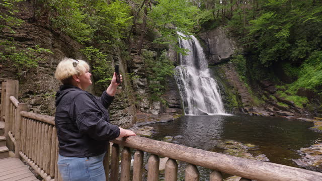 Bushkill Falls, PA is popular place for tourists. Mature woman photographs waterfall from wooden walkway in the forest