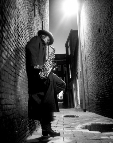 saxophone player standing on one leg against wall in poorly illuminated alley playing on the saxophone