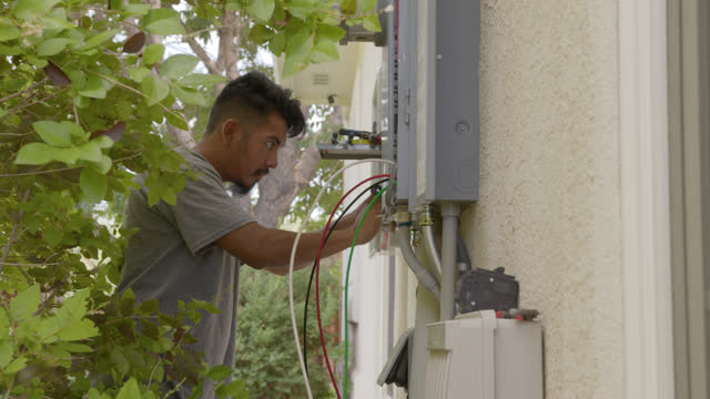 Wide View of a Latino Man Installing Wiring a Circuit Breaker Disconnect Box for a DIY Solar Installation on a Suburban Home in the USA