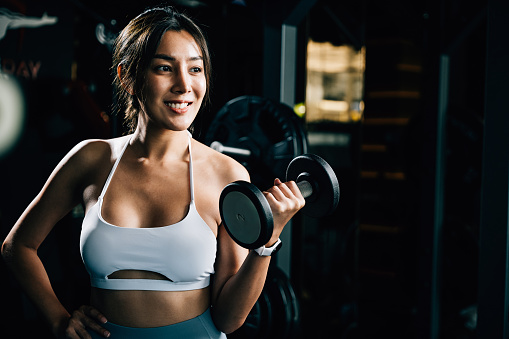 A young and beautiful girl in gym clothes, holding a dumbbell and lifting weights, showcases her strength and fitness for a healthy lifestyle.