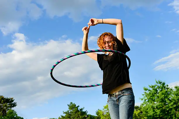 A red headed female outside playing with a hula hoop.  Sky, clouds and trees in the background.