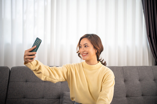 An Asian girl with a big smile sits in the living room. She has fun taking cheerful use smartphone video conference selfies and looking at the camera