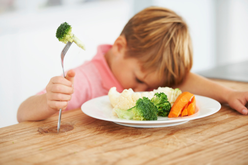 A cute young boy with his head on the table while holding a piece of broccoli on his fork