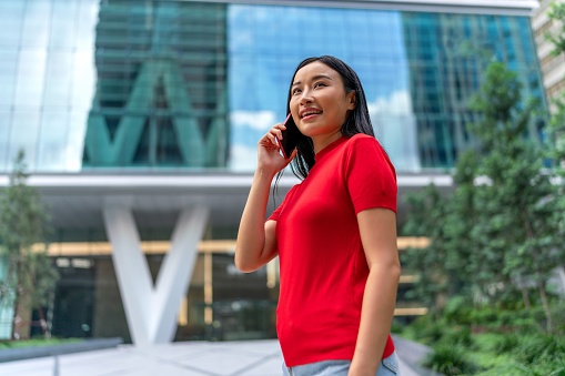 With a mobile phone in hand, the young Asian woman stays connected while on the move, seamlessly engaging with on-the-move modern activities like social media and messaging as well as exemplifying efficiency through digital banking and modern professionalism.