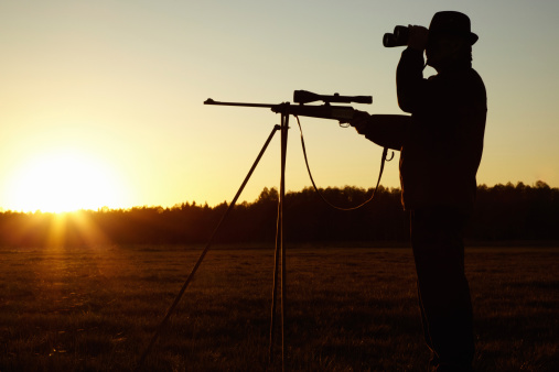 A silhouette of a man in the wildlife with his sniper rifle ready and looking through his binoculars