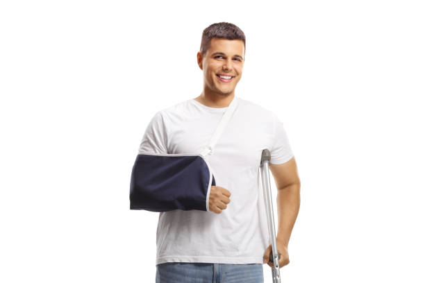 young man with a broken arm wearing an arm splint and standing with a crutch - physical injury men orthopedic equipment isolated on white imagens e fotografias de stock