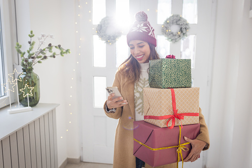 Young woman entering at home for Christmas. She is holding gift boxes and using smart phone.