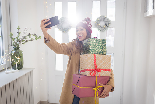 Young woman entering at home for Christmas. She is holding gift boxes and making selfie.
