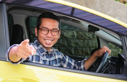 Adult Asian man smiling and pointing at the camera from inside a car