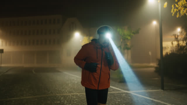 SLO MO Sportsman with Glowing Headlamp Putting On Wireless Headphones and Jogging on Street at Night