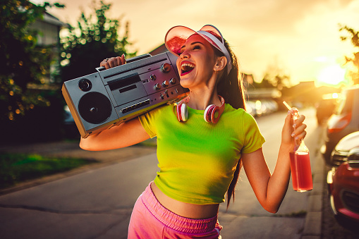 Beautiful young woman in neon clothes drinking colorful juice on the street and carrying a boombox radio on her shoulder.