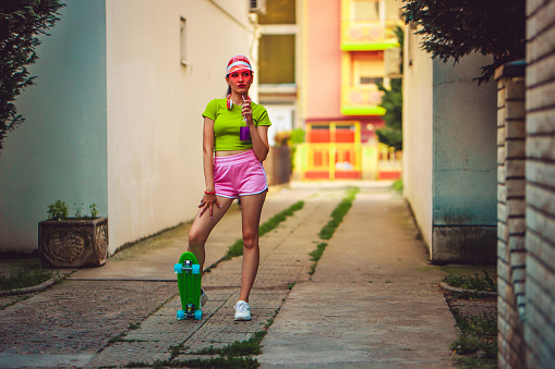 Beautiful young skateboarder woman in neon clothes drinking colorful juice on the street.