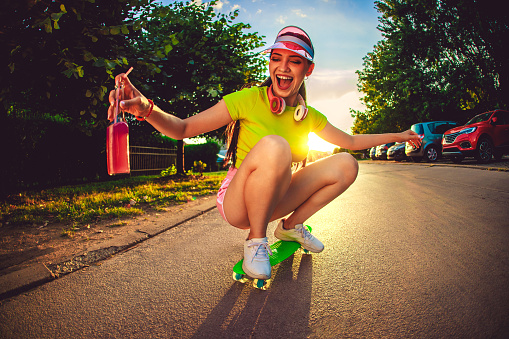 Beautiful young skateboarder woman in neon clothes drinking colorful juice and riding a skateboard on the street.