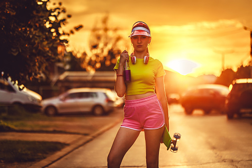 Portrait of a beautiful young skateboarder woman in neon clothes drinking colorful juice on the street.