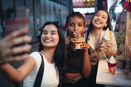 Joyful female friends are taking selfies with ice cream in the city at night.