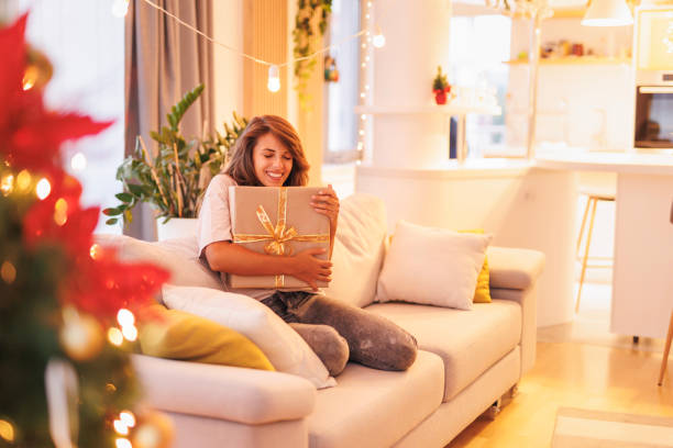 Woman hugging Christmas gift box while relaxing at home stock photo