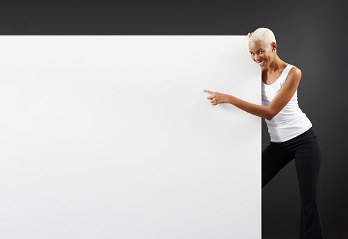 Smiling black woman pointing at a white blank advertising banner sign with copy space. A store and mall advertising billboard for promotional event or offers. Black Friday shopping concept