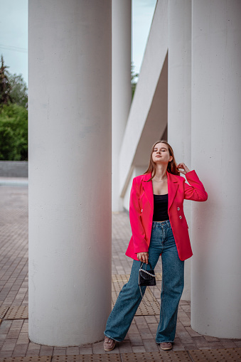 a young business girl in a bright fuchsia jacket and jeans stands walking through columns on the street in summer