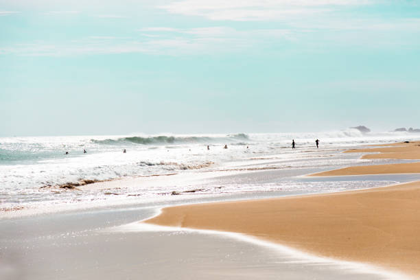 A stunning golden beach with big turquoise waves crashing onto the smooth sand. The beach is wide and the sand is soft. In the distance, there are a few people enjoying the water and the view. The sky is blue and the horizon is visible. The photo is taken from a low angle, giving a sense of the vastness of the beach.