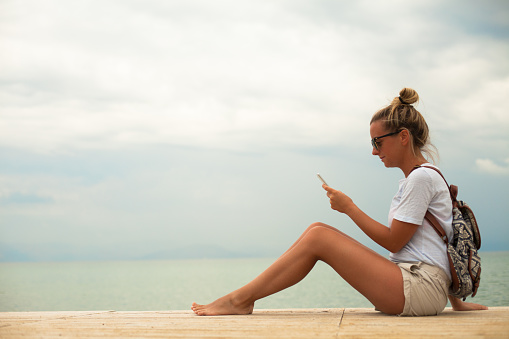 Young woman texting on her phone, sitting on the coast and relaxing.