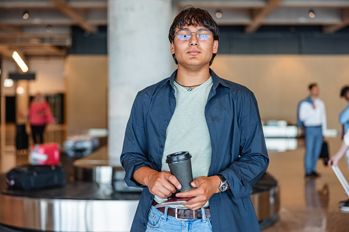 Three-quarter front shot of a young mixed-race man looking a little serious at the camera while holding a cup of coffee and his documents at the airport arrivals zone.