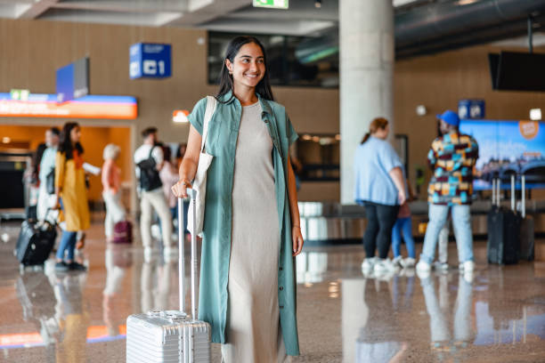 Pretty Mixed-Race Woman Pulling Her Gray Suitcase At The Airport Three-quarter side shot with blurred background of a casually dressed attractive young woman pulling her gray suitcase from the baggage claim area to the exit of the airport, surrounded by passengers. walking into glass door stock pictures, royalty-free photos & images