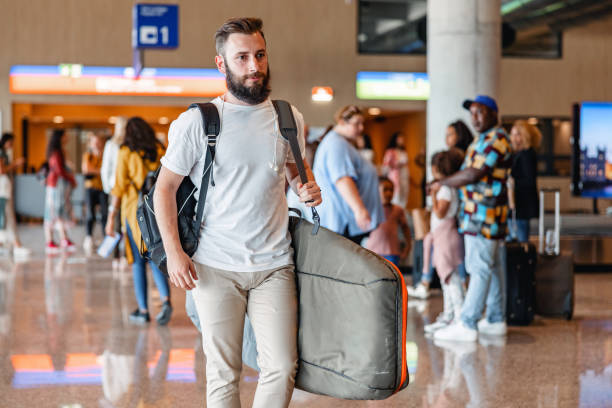 Sporty Caucasian Man Carrying A Surfboard At The Airport Three-quarter side shot with blurred background of a Caucasian mid-adult man carrying a surfboard and a backpack while walking through the airport arrival zone during the day. walking into glass door stock pictures, royalty-free photos & images