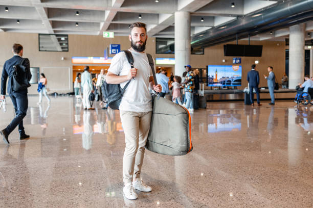 Mid-Adult Man Looking At Camera While Walking At The Airport Full length front shot with blurred background of a Caucasian mid-adult man carrying a surfboard in a gray case and a black backpack while looking at the camera at the airport arrival zone during the day. walking into glass door stock pictures, royalty-free photos & images