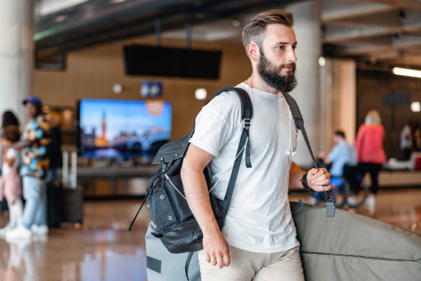 Mid-Adult Man Walking Through The Airport With Baggage Three-quarter side shot with blurred background of a Caucasian mid-adult man carrying a surfboard and a backpack while walking through the airport arrival zone during the day. walking into glass door stock pictures, royalty-free photos & images