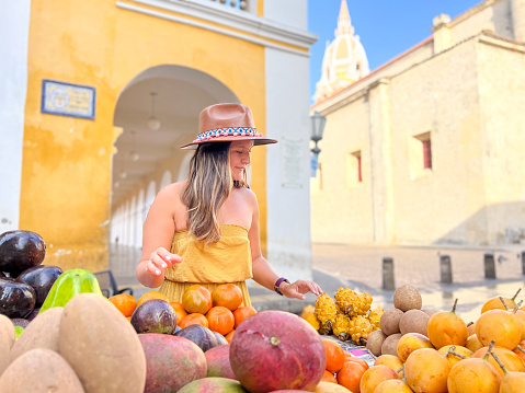She smiles, picking up and looking at the beautiful tropical fruits from the market stall, low angle view. Sunny Cartagena cityscape on the background