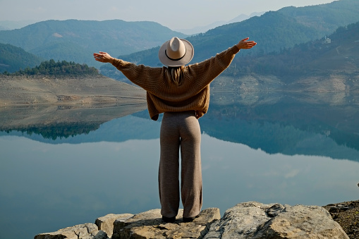 Female traveler wearing a broad brim hat enjoying beautiful view of a mountain lake. Young woman experiencing spiritual uplifting from being alone in nature. Background, copy space, back view.