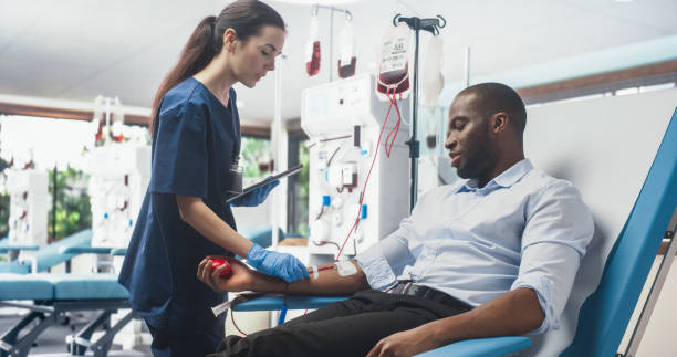 un homme d’affaires noir donne du sang pour des personnes dans le besoin dans un hôpital lumineux. infirmière avec tablette venant vérifier les progrès et le bien-être du donneur. don pour les patients en chirurgie cardiaque. - don du sang photos et images de collection