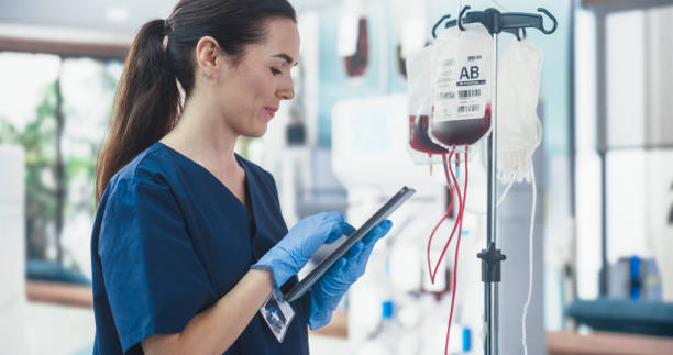 female nurse holding tablet computer and checking the blood bag in donation center. professional caucasian woman observing donation process in bright hospital. healthcare and charity concept. - bloedbank stockfoto's en -beelden