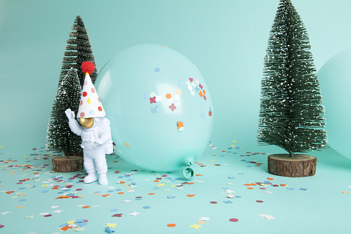 a cosmonaut exploring a snowy forest like a new planet. They are surrounded by confetti, balloons and christmas tree. The composition is in turquoise-green color palette. Minimalist, trendy still life photography.