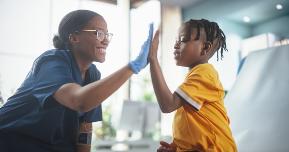 Young African American Boy Sitting In The Chair In Bright Hospital And Getting His Flu Vaccine. Female Black Nurse Is Finished Performing Injection. Professional Woman High-Fives A Kid For Being Brave
