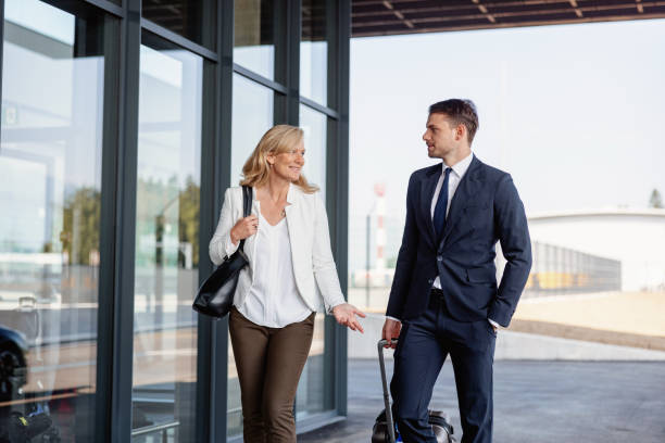 Caucasian Colleagues Of Different Ages About To Enter The Airport Three-quarter shot with blurred background of two Caucasian colleagues of different ages discussing some meeting details while walking towards the entrance of the airport to check-in for their upcoming business trip on a sunny day. walking into glass door stock pictures, royalty-free photos & images