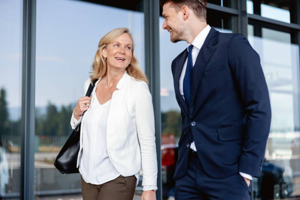 Caucasian Colleagues Of Different Ages Walking Towards The Airport Entrance Three-quarter shot with blurred background of two Caucasian colleagues of different ages discussing some meeting details while walking towards the entrance of the airport to check-in for their upcoming business trip on a sunny day. walking into glass door stock pictures, royalty-free photos & images