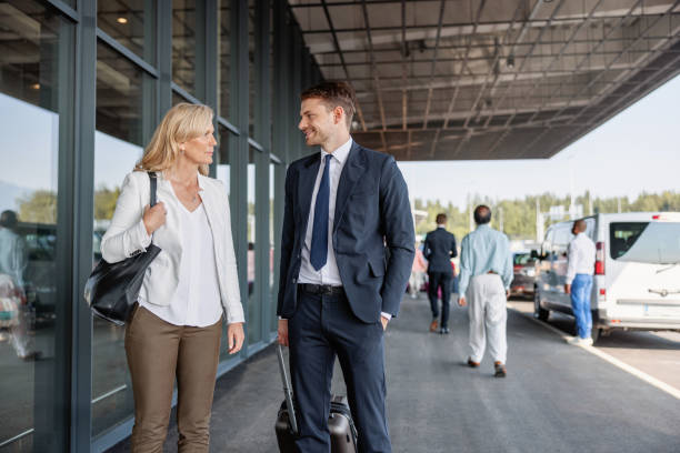 Caucasian Colleagues Of Different Ages Walking Towards The Airport Entrance Three-quarter shot with blurred background of two Caucasian colleagues of different ages discussing some meeting details while walking towards the entrance of the airport to check-in for their upcoming business trip on a sunny day. walking into glass door stock pictures, royalty-free photos & images