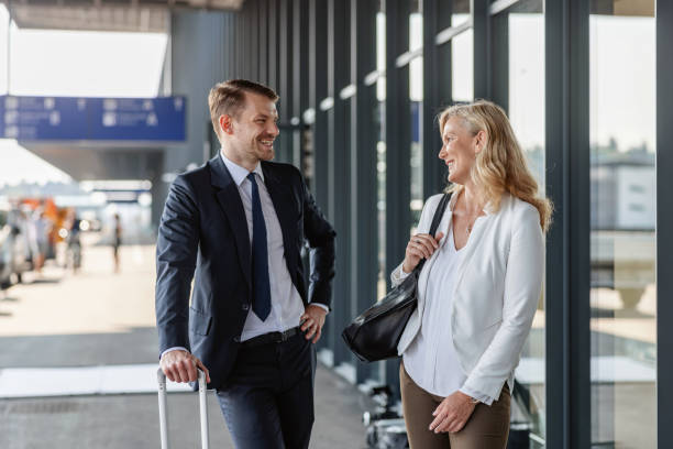 Caucasian Colleagues Of Different Ages Walking Towards The Airport Entrance Three-quarter shot with blurred background of two Caucasian colleagues of different ages smiling and talking about some meeting details while walking towards the entrance of the airport to check-in for their upcoming business trip on a sunny day. walking into glass door stock pictures, royalty-free photos & images