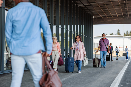 Full length side shot with blurred motion of a group of passengers of different ethnic groups and ages walking towards the airport or leaving it on a bright day.