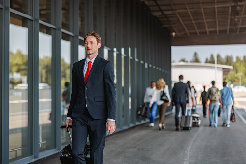 Three-quarter side shot with blurred background of a good looking Caucasian airplane pilot walking towards the airport entrance pulling a black suitcase and wearing a formal dark suit and a red tie as a part of his work uniform surrounded by many passengers.