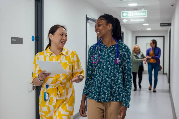 General Practitioners in Hospital Walkway Front view three quarter length of two doctors having a conversation in a hospital corridor, wearing smart casual clothing. They discuss patient care, reflecting their dedication and expertise. The scene exudes professionalism and trust in their collaborative approach to medicine in a hospital in Newcastle, England. Behind them a mid adult woman is walking down the corridor with her senior mother. hospital card stock pictures, royalty-free photos & images