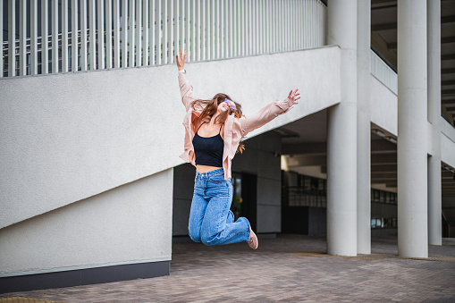 a young girl with long hair, wearing glasses and jeans, joyfully jumps in the air in the city against the backdrop of a building with white columns in the summer