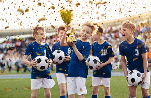 Group of school boys celebrating together the winning of a competition running on a soccer field in confetti. Soccer team rising golden cup trophy after winning youth tournament