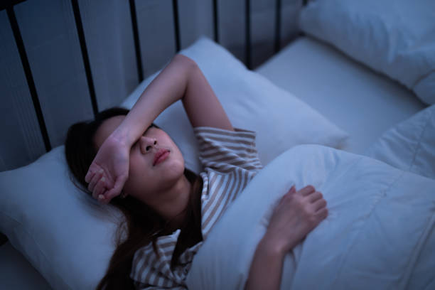 真夜中に寝室のアパートで仕事の疲労と悪夢に苦しんでいるため、腕から目を覆いながら一人で眠ろうとしているアジアの不眠不休の女性 - 不眠症 ストックフォトと画像