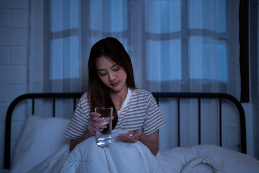 Asian insomnia girl decide take a sleeping pill and drink water in a glass before fall asleep from exhaustion while wearing casual pajama and sitting in bed at night alone in the house