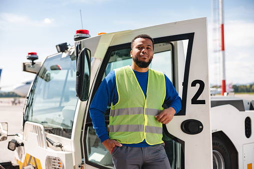Three quarter length photo of a Hispanic male airport worker wearing a reflective vest leaning on a pushback tug - towing vehicle used at the airports.