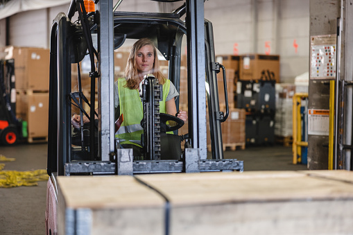 Caucasian female airport warehouse worker driving a forklift and moving parcels around.