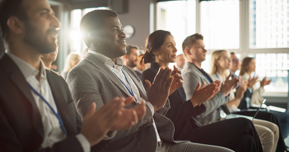 African American Man Sitting in Crowded Auditorium at a Tech Conference. Black Businessman Listening To Keynote. Specialist Watching Innovative Technology Presentation And Clapping With Audience.